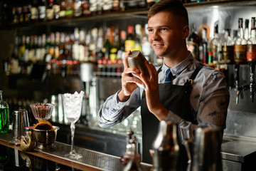 young smiling bartender standing at bar counter with shaker in his hands.