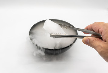 Hand holding ice tweezer with dry ice over black bowl of smoky white in motion isolated on white