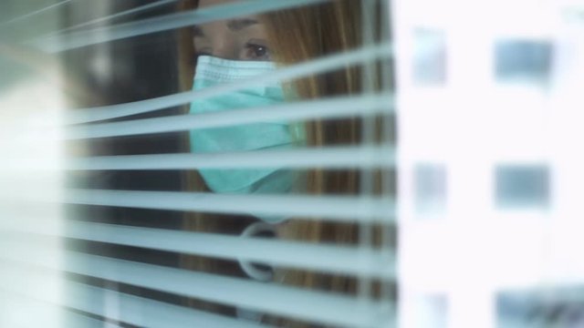 Sad young woman wearing medical protection mask looking out the window through the blinds at home. Home quarantine coronavirus. Social distancing, self isolation.