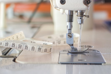 Needle presser foot with blue thread of white industrial sewing machine use sew cloth and tape measure close-up.