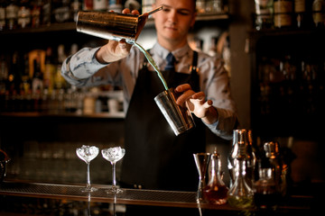 young barman pours cocktail in shaker. Two glasses with ice stand on bar.