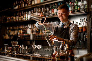 young smiling barman pours cocktail in shaker. Two glasses with ice stand on bar.