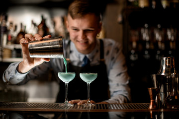 young smiling bartender pours cocktail from shaker into glasses