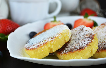 Fried cottage cheese pancakes or syrniki with fresh berries on a white plate on the black table. Gluten free. Traditional breakfast of Ukrainian and Russian cuisine. Horizontal, close up.