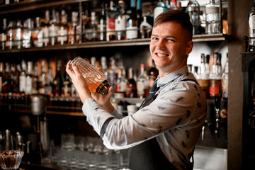 young smiling bartender at bar mixes cocktail in a shaker.