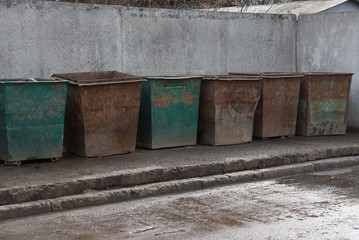 a row of old iron rusty garbage cans stand against a gray concrete wall on asphalt on the street