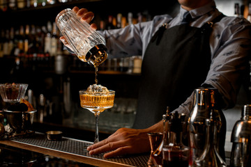 Professional bartender in black apron pours drink from shaker into glass. - obrazy, fototapety, plakaty