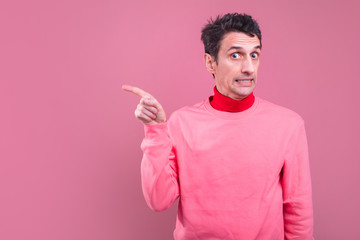 Picture of worried young man point with figer to left and look on camera. Guy with strange weird face reaction. Isolated over pink background.