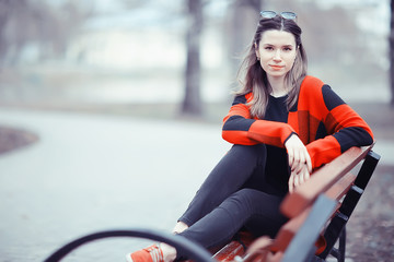 Naklejka premium girl is sitting on a garden bench / beautiful model posing sitting in coat on bench in city park, beautiful girl