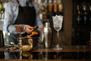 Beautiful view of bar on which stands glass with ice, shaker and vase with slices of fruits.