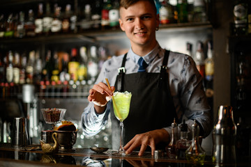Young smiling barman decorates cocktail with ice in glass by two sticks.