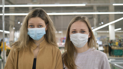 Two young women in medical masks stand sad in a supermarket and look at the camera. Protection from the coronavirus pandemic, preparation for quarantine.