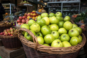 Green apples in wicker basket at a market for sale. In the background are counters with seasonal vegetables.