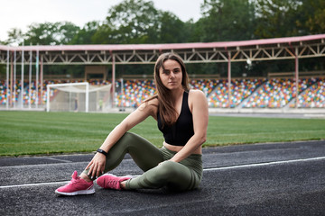 Young brunette woman, wearing black top and green leggings, sitting on city stadium on summer morning, resting relaxing after training. Portrait of sporty girl, posing. Healthy active life concept