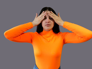 Young beautiful girl with dark hair in a bright orange sweater on a gray background. She covers her face with her own hands