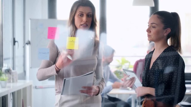 Video of two business young women working together on wall glass with post it stickers on coworking space.