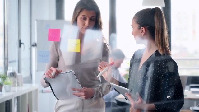 Video of two business young women working together on wall glass with post it stickers on coworking space.