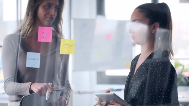 Video of two business young women working together on wall glass with post it stickers on coworking space.