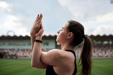 Young brunette sportswoman, wearing black top and green leggings, doing yoga pose on city stadium on summer morning, Stretching training outside on pink yoga mat. Healthy active life concept.