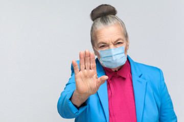 aged grandma with surgical medical mask showing stop sign at camera. Grandmother with light blue suit and pink shirt standing with collected bun hair. indoor studio shot, isolated on gray background