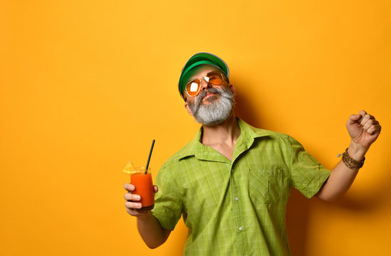 Bearded Man In Green Sun Visor, Shirt And Sunglasses. Holding Glass Of Squeezed Juice, Clenched Fist, Posing On Orange Background