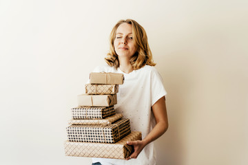 Young charming cheerful caucasian blonde holds a stack of gift boxes wrapped in craft paper....