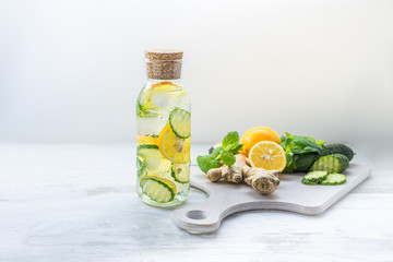Detox Sassy water with lemon, cucumber, mint. A glass bottle of clean, cool and fresh drink stands on a white wooden table with ingredients. Strengthening immunity, diet. Still life with copy space.