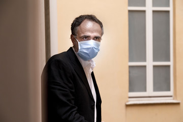 Fototapeta na wymiar Manager con mascherina chirurgica si affaccia al balcone con aria sconsolata, sullo sfondo la finestra di un edficio