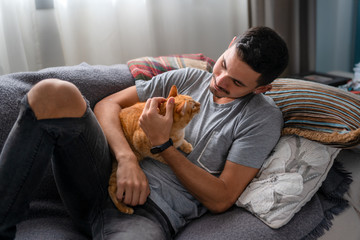 hombre joven recostado en un sofa abraza a un gato atigrado y sonrie