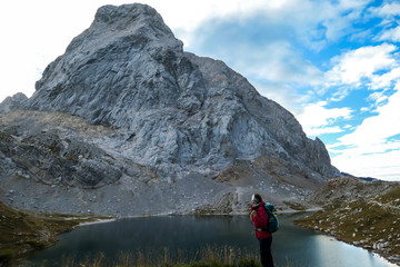 Naklejka premium A woman in hiking outfit standing by the Wolayer Lake in Austrian Alps. There is massive, rocky mountain on the other side of the lake. New day beginning. Soft reflections in the lake. Happiness