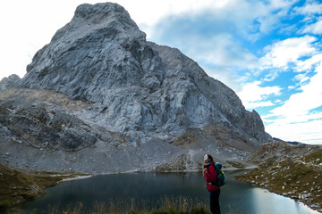 Fototapeta premium A woman in hiking outfit standing by the Wolayer Lake in Austrian Alps. There is massive, rocky mountain on the other side of the lake. New day beginning. Soft reflections in the lake. Happiness
