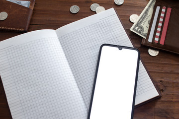 A smartphone with a white screen on top of an open notebook next to dollars and rubles on a wooden table next to a leather holder and wallet.