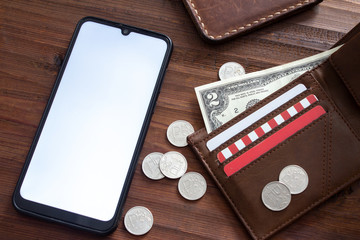 A wallet in which dollar and rubles are next to a smartphone which has a white screen on a wooden background.