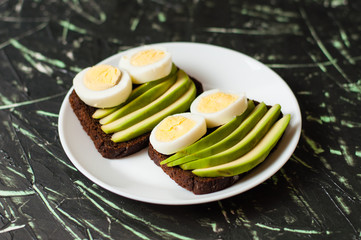 Sandwiches with brown bread, avocado and egg on a dark background, top view