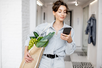 Portrait of a young and cheerful woman standing with mobile phone and shopping bag full of fresh food at home. Concept of buying products online - obrazy, fototapety, plakaty