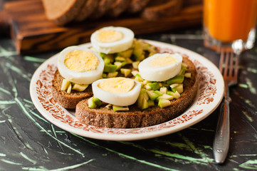 A glass of juice, sandwiches with avocado and egg, black whole grain bread.