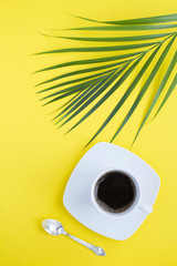Black coffee in the white cup and palm leaves on the yellow  background. Location vertical. Top view. Copy space.