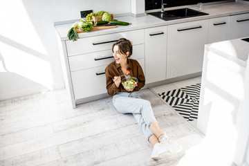 Young woman sitting on the white kitchen floor, eating salad while relaxing at home. Healthy eating, food and lifestyle concept