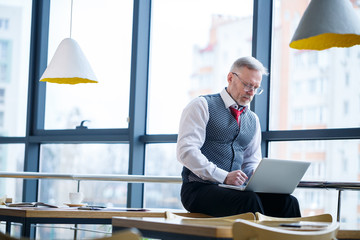 Fototapeta premium Adult male businessman, teacher, mentor working on a new project. Sits by a large window on the table. He looks at the laptop screen.
