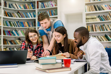 Four positive smiling multiethnic boys and girls sitting at the table in modern college library and jointly doing home task or searching information for learning subject