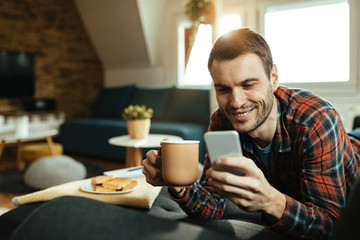 Happy man drinking coffee and text messaging on mobile phone at home.