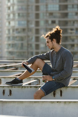 Fototapeta na wymiar Man sitting on roof and reading book, drinking coffee, tea, drink in paper cup in grey shirt, blue jeans shorts, curly hair collected in high ponytale . Background cityscapes. Urban lifestyle concept