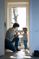 Man putting a medical mask on little girl's face. Father and daughter with breathing masks near the door.