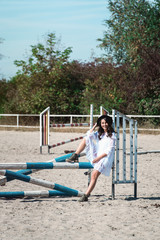Fototapeta premium Beautiful sexy girl posing on the horizontal crosspiece on a horse epodrom. Female wearing white dress, black hat and black boots