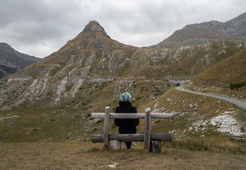 Young woman with blue hair in a black coat sits on a wooden bench on a mountainside