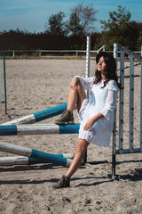 Beautiful sexy girl posing on the horizontal crosspiece on a horse epodrom. Female wearing white dress, black hat and black boots