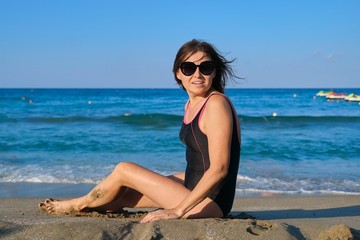 Healthy beautiful middle-aged woman in swimsuit sunglasses sitting on seashore