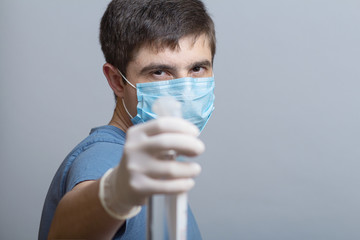 young man in surgical mask holding up bottle of disinfectant spray on a gray studio background,...