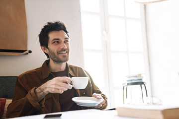 Image of joyful young man smiling and drinking tea while resting