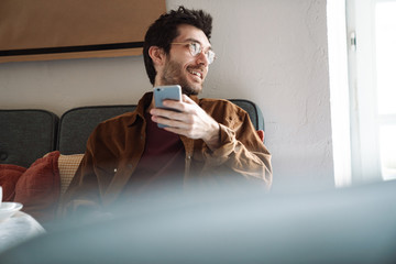 Image of handsome joyful young man wearing eyeglasses using cellphone